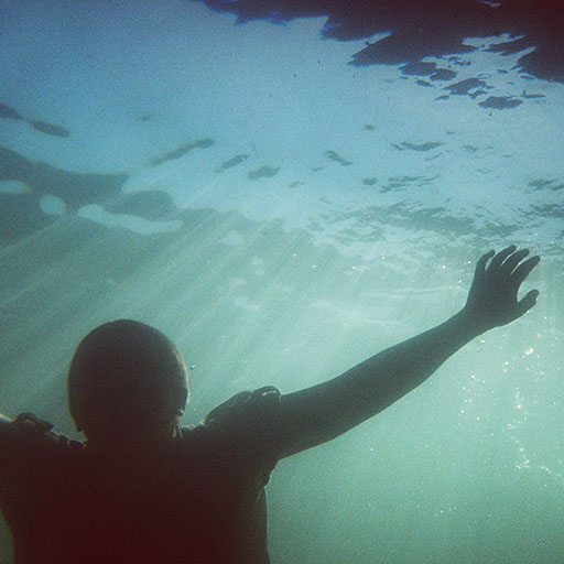 Kelpie, shaved head and clothed in a short sleeved linen shirt, floats underwater with one arm thrown out to the side. The atmosphere is mysterious.